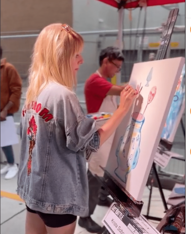 a person with long blond hair wearing a denim jacked painting at a canvas